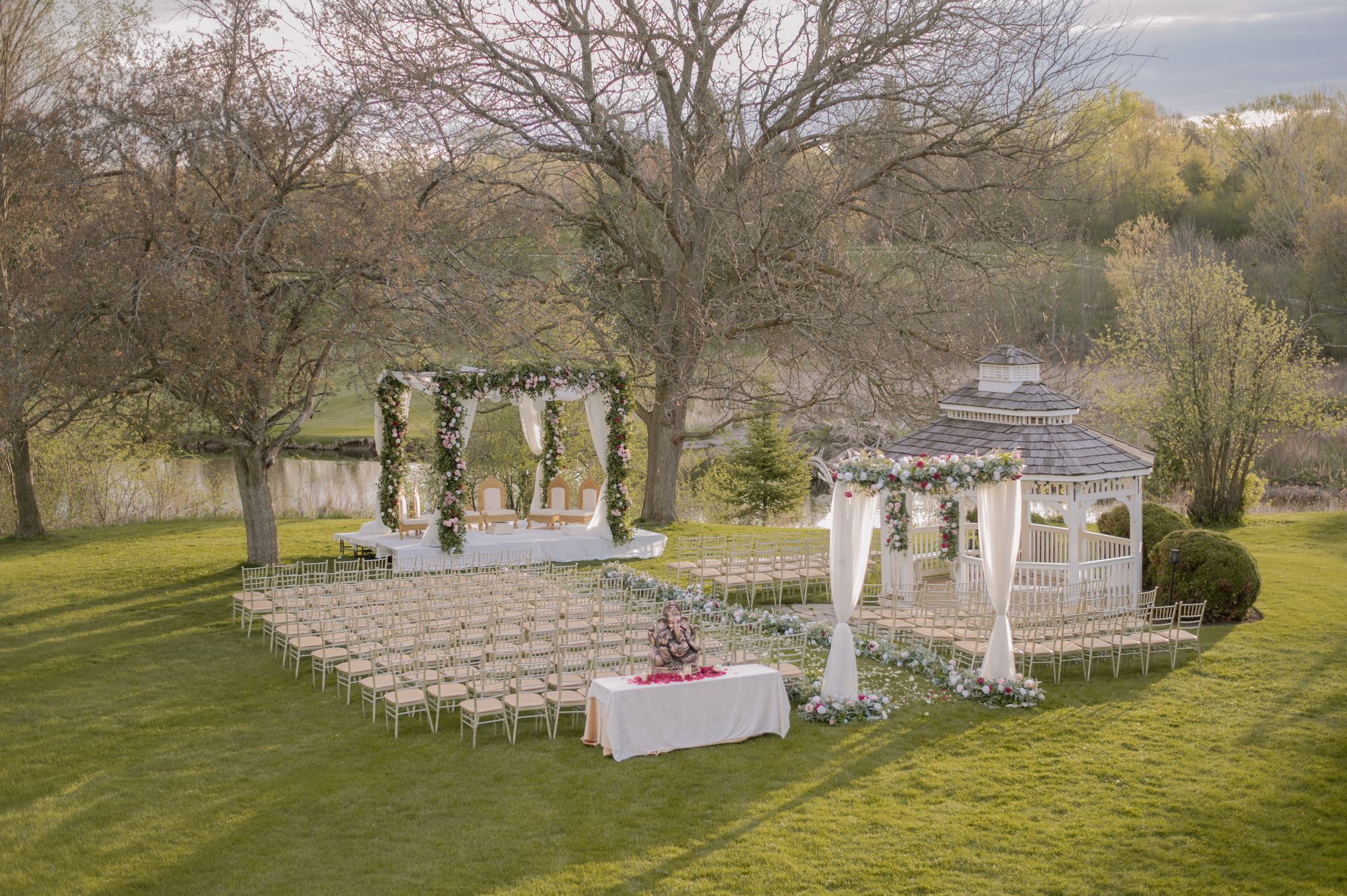 An outdoor mandap and floral gazebo set for a wedding ceremony beside a pond at golden hour